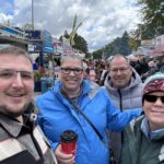 Four smiling adults in jackets stand close together at an outdoor fair, with food stands, signs, and a crowd in the background. The sky is cloudy, and one person holds a red coffee cup.