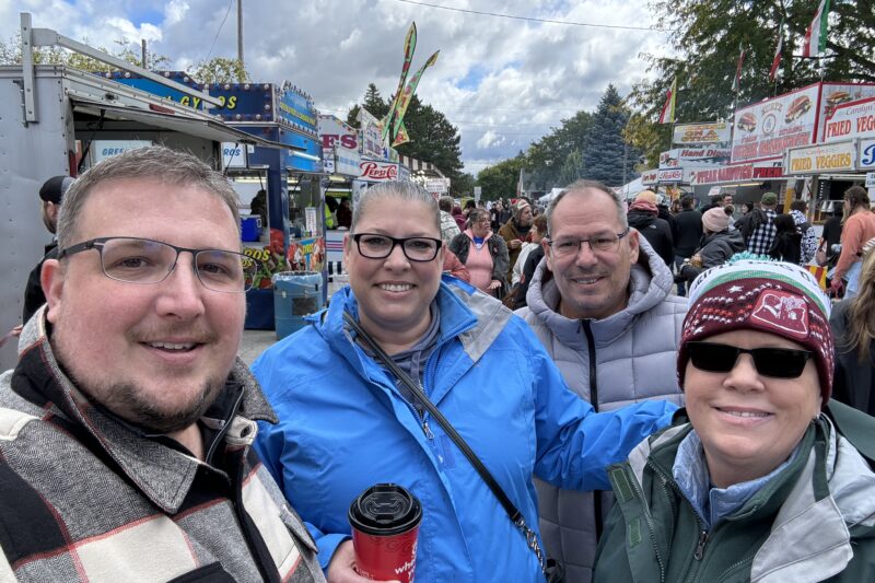 Four smiling adults in jackets stand close together at an outdoor fair, with food stands, signs, and a crowd in the background. The sky is cloudy, and one person holds a red coffee cup.