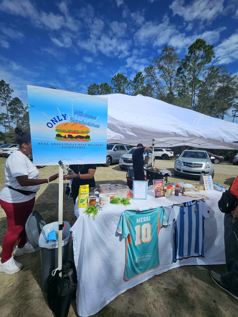 Outdoor food vendor booth serving Argentine-style street food with a sign, table setup, and customers browsing