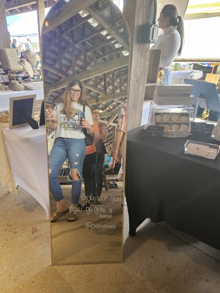 A woman takes a mirror selfie at a market stall, smiling and holding up a peace sign. The mirror reads, You look good. Now go grab a treat. @dijondrizzle. Baked goods and displays are visible nearby.