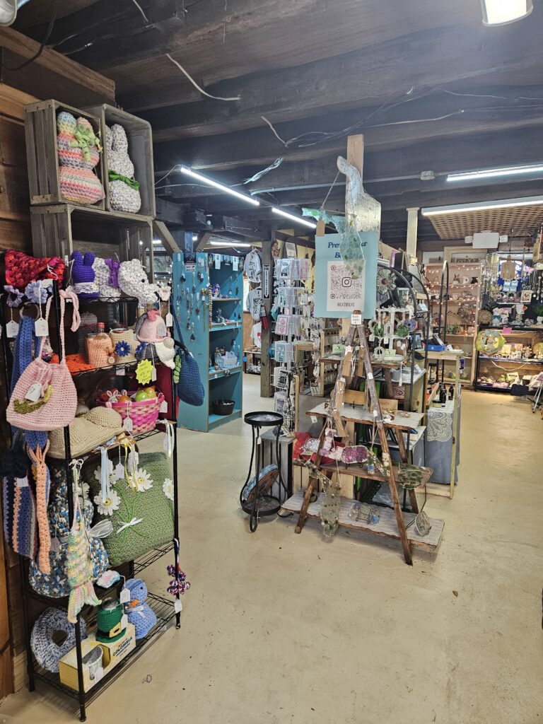 Craft shop interior with shelves displaying crocheted bags and dolls on the left, a teal shelf with various items in the center, and more handmade crafts and decor on tables and shelves throughout the cozy, well-lit space.