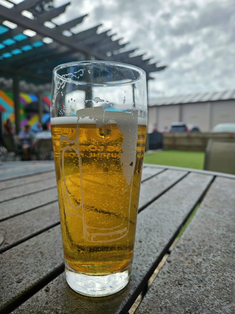A nearly empty glass of beer sits on a wooden outdoor table, with foam residue inside the glass. In the background are blurred people, a rainbow mural, and a partly cloudy sky.