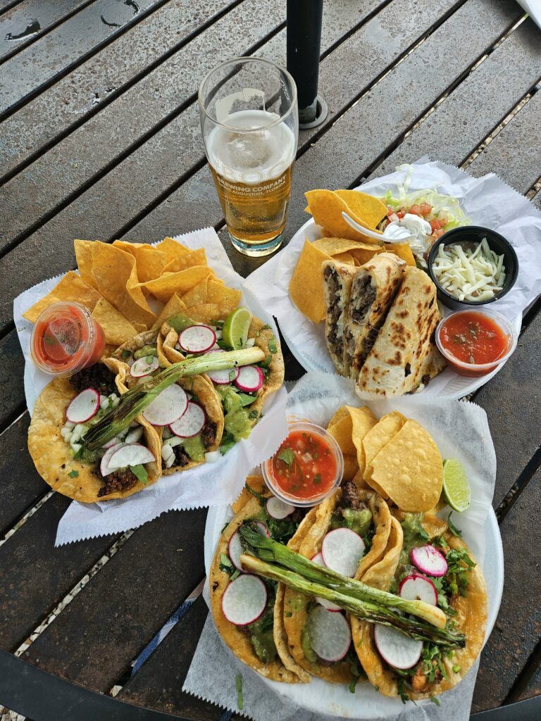 Two plates of street tacos topped with grilled asparagus, radish slices, and cilantro, served with tortilla chips, salsa, and lime. A side of quesadillas with veggies and a half-full pint of beer are on a rustic wooden table.