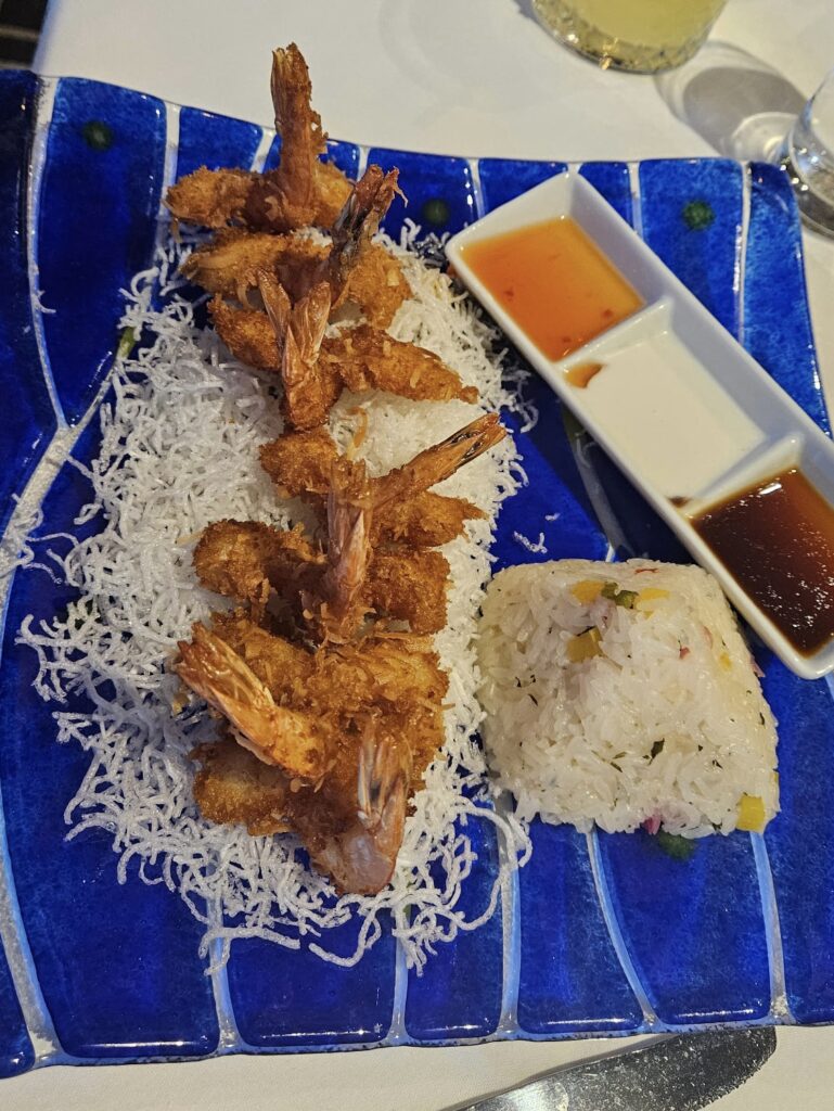 A blue plate with crispy coconut fried shrimp on a bed of shredded white noodles, a serving of mixed vegetable rice, and two small dishes of dipping sauces—one orange, one brown.