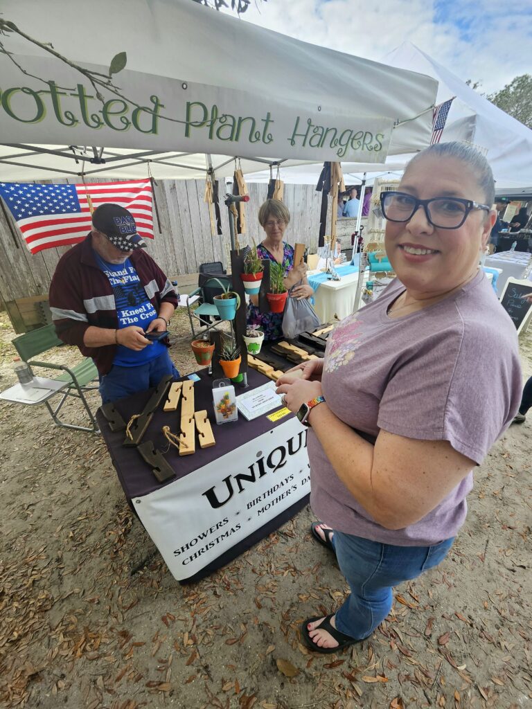 Vendor selling potted plant hangers at an outdoor market booth with American flags and handmade items on display
