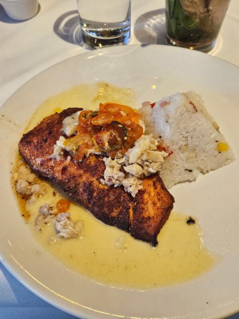 A plate with a seasoned, cooked fish fillet topped with a creamy sauce and sautéed vegetables, served alongside a molded portion of white rice mixed with small pieces of vegetables. A glass of water is in the background.