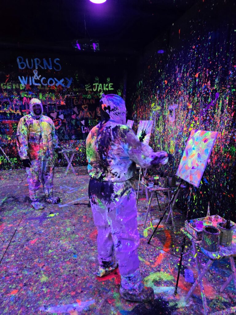Two people in paint-splattered white suits create art in a dark room covered with neon paint splatters and graffiti, illuminated by blacklight. One paints at an easel, while the other observes.