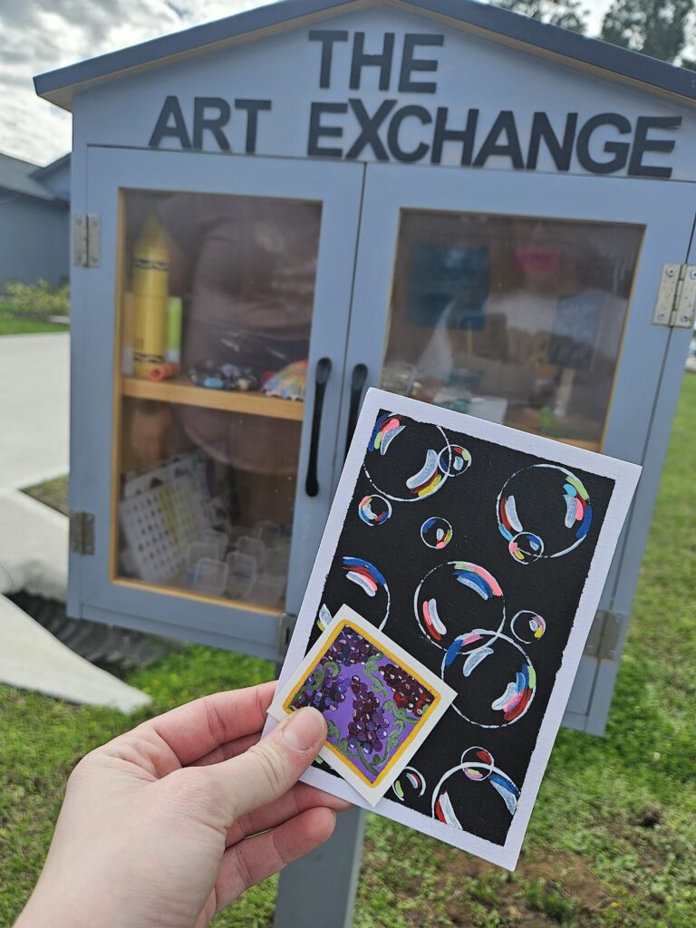 A hand holds two colorful art pieces in front of a small outdoor cabinet labeled THE ART EXCHANGE, which contains various art supplies and items for swapping. Grass and a sidewalk are visible in the background.