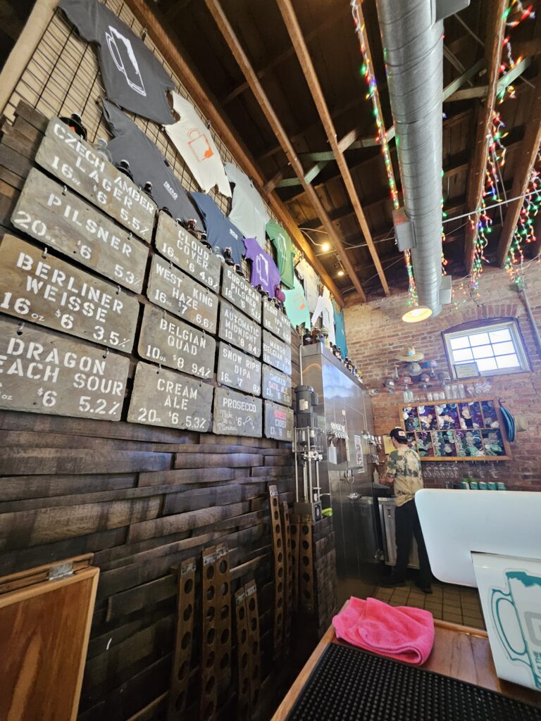 A cozy bar with a rustic interior, featuring a wooden wall displaying beer options on boards, T-shirts hanging above, exposed pipes, string lights, and a person standing near drink taps.