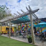 People dine outdoors under a wooden pergola with a blue-tinted roof. Colorful mural on the wall and a yellow food truck are in the background. Green artificial grass and umbrellas add to the vibrant, relaxed setting.