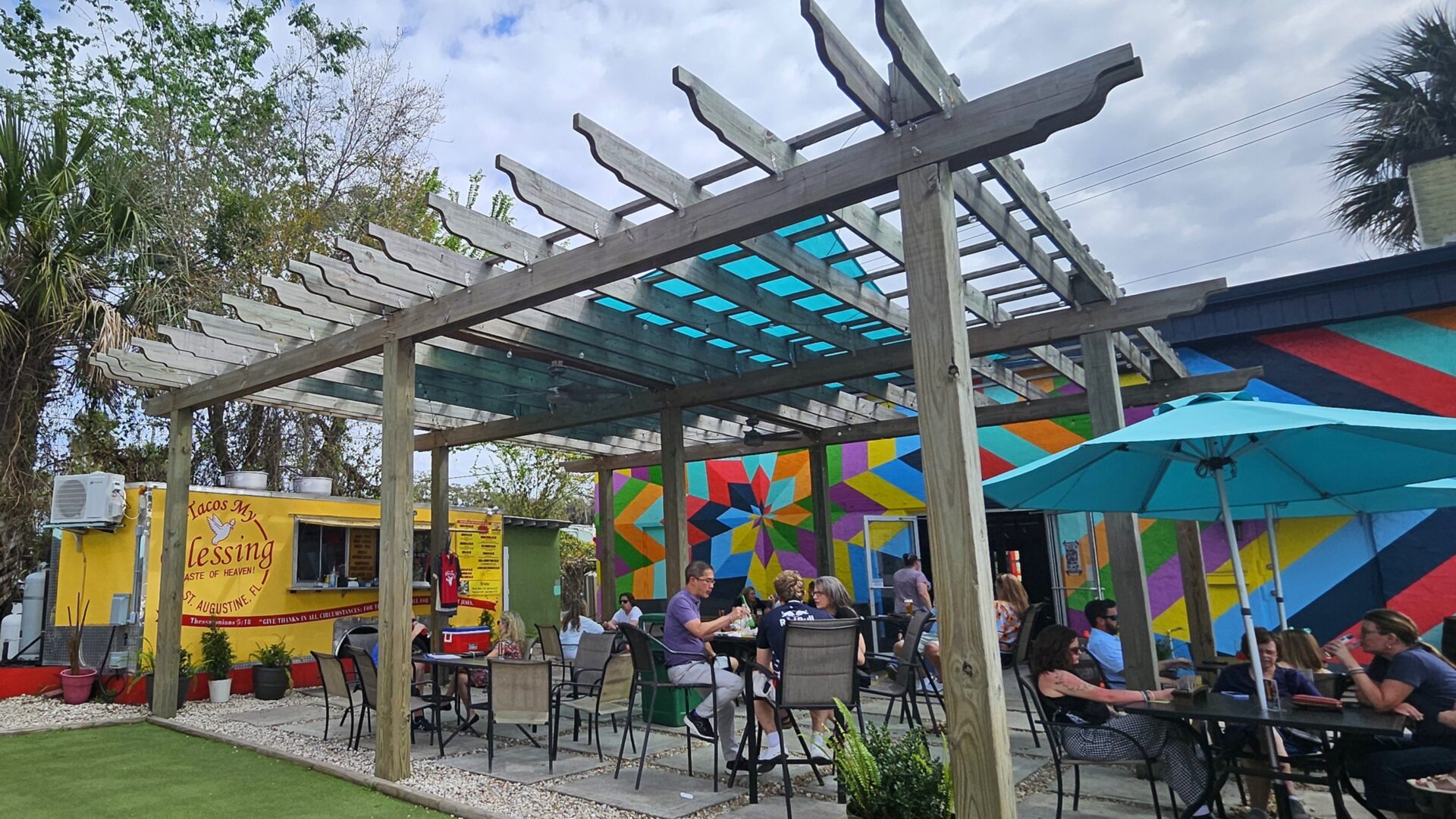 People dine outdoors under a wooden pergola with a blue-tinted roof. Colorful mural on the wall and a yellow food truck are in the background. Green artificial grass and umbrellas add to the vibrant, relaxed setting.