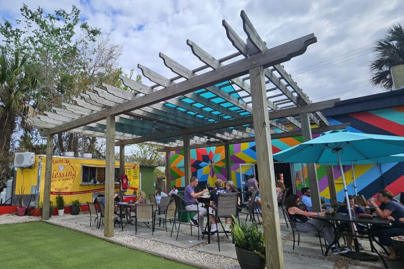 People dine outdoors under a wooden pergola with a blue-tinted roof. Colorful mural on the wall and a yellow food truck are in the background. Green artificial grass and umbrellas add to the vibrant, relaxed setting.