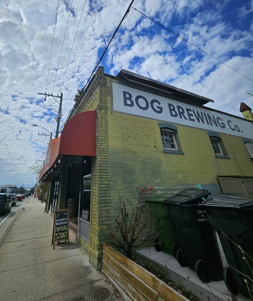 A yellow brick building with a sign reading BOG BREWING CO. stands on a street corner under a partly cloudy sky. A sandwich board advertises imperial stout near the entrance; trash bins are beside the building.
