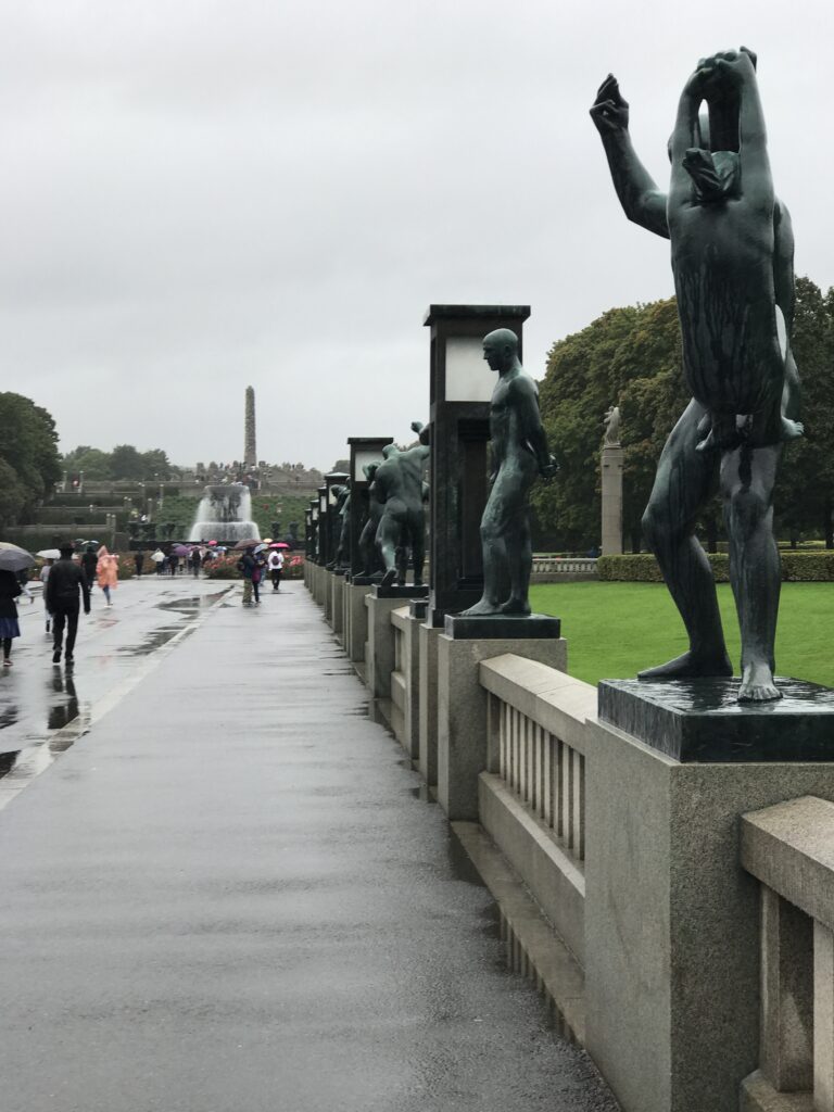 Walkway lined with bronze sculptures in Vigeland Park with visitors walking in the rain