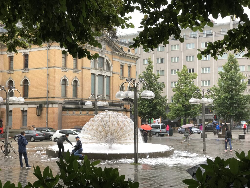 Fountain in Oslo city square with pedestrians walking in the rain