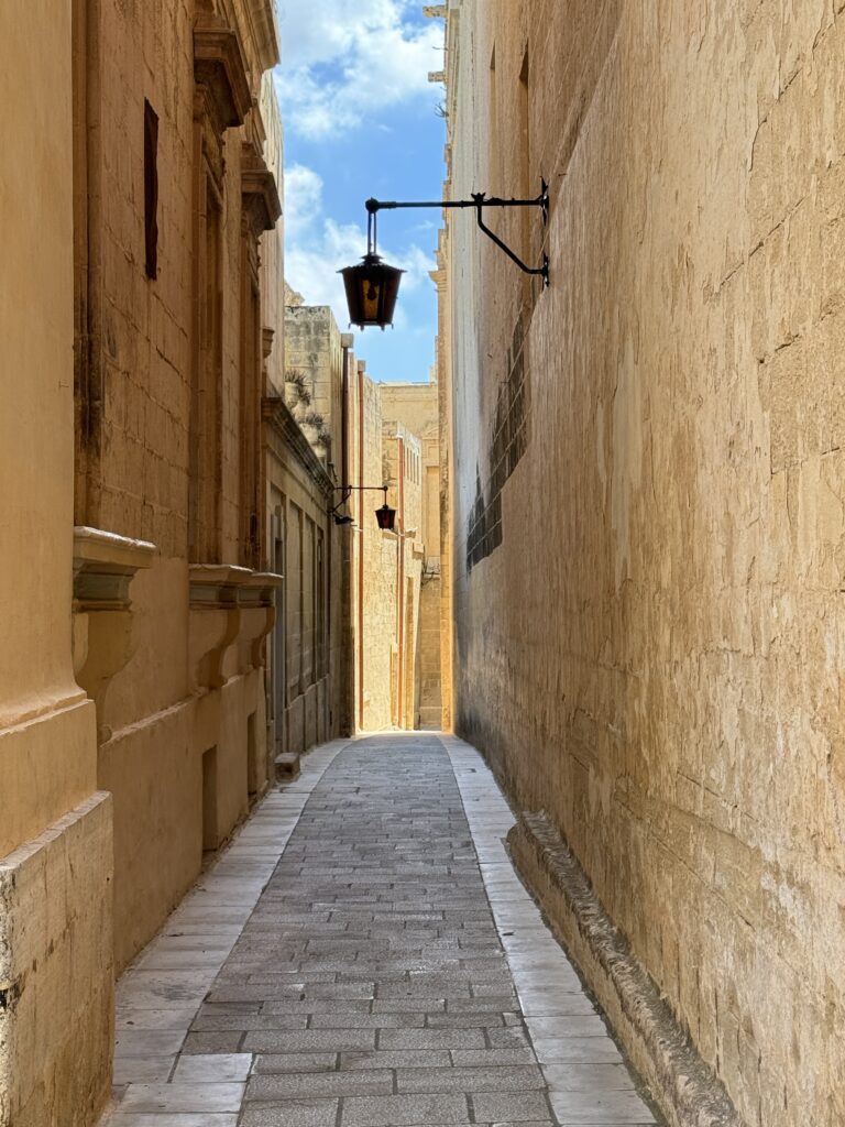 Quiet, narrow street in Mdina lined with tall limestone walls and a hanging lantern, leading toward sunlight at the end.