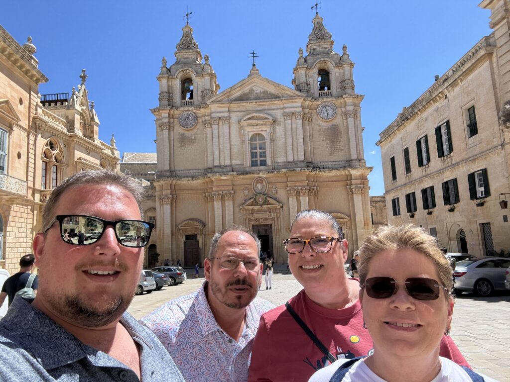 Group of travelers posing in front of a historic cathedral in Mdina, Malta, under a bright blue sky.