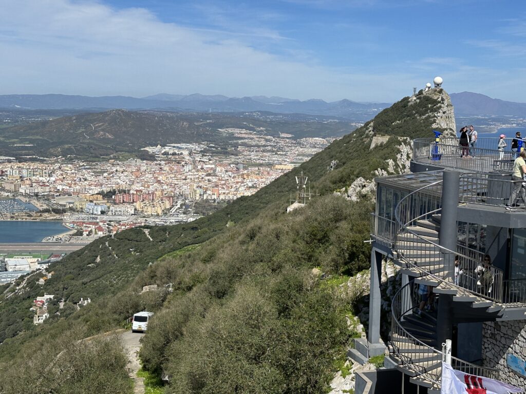 View from the Rock of Gibraltar overlooking the city, marina, and coastline below, with a scenic viewing platform and visitors standing along the edge of the mountainside.