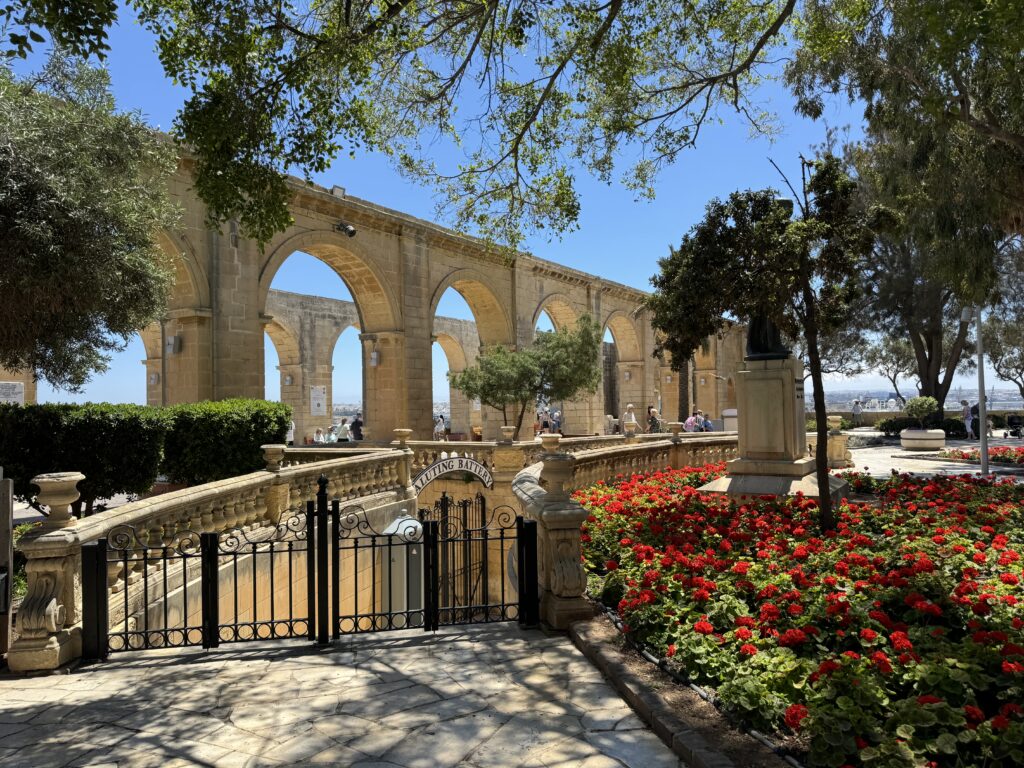 Upper Barrakka Gardens in Valletta, Malta, with stone arches, shaded walkways, and vibrant red flowers overlooking the harbor.