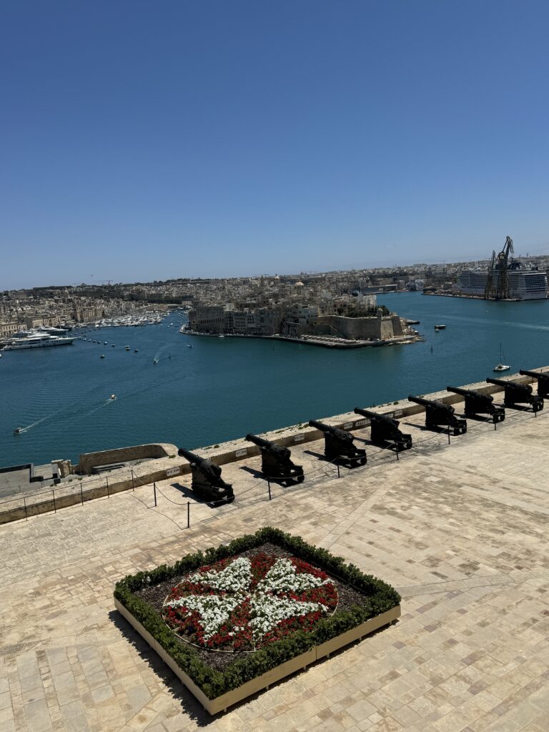 Row of historic cannons at the Saluting Battery in Valletta overlooking the Grand Harbour with boats and city views across the water.