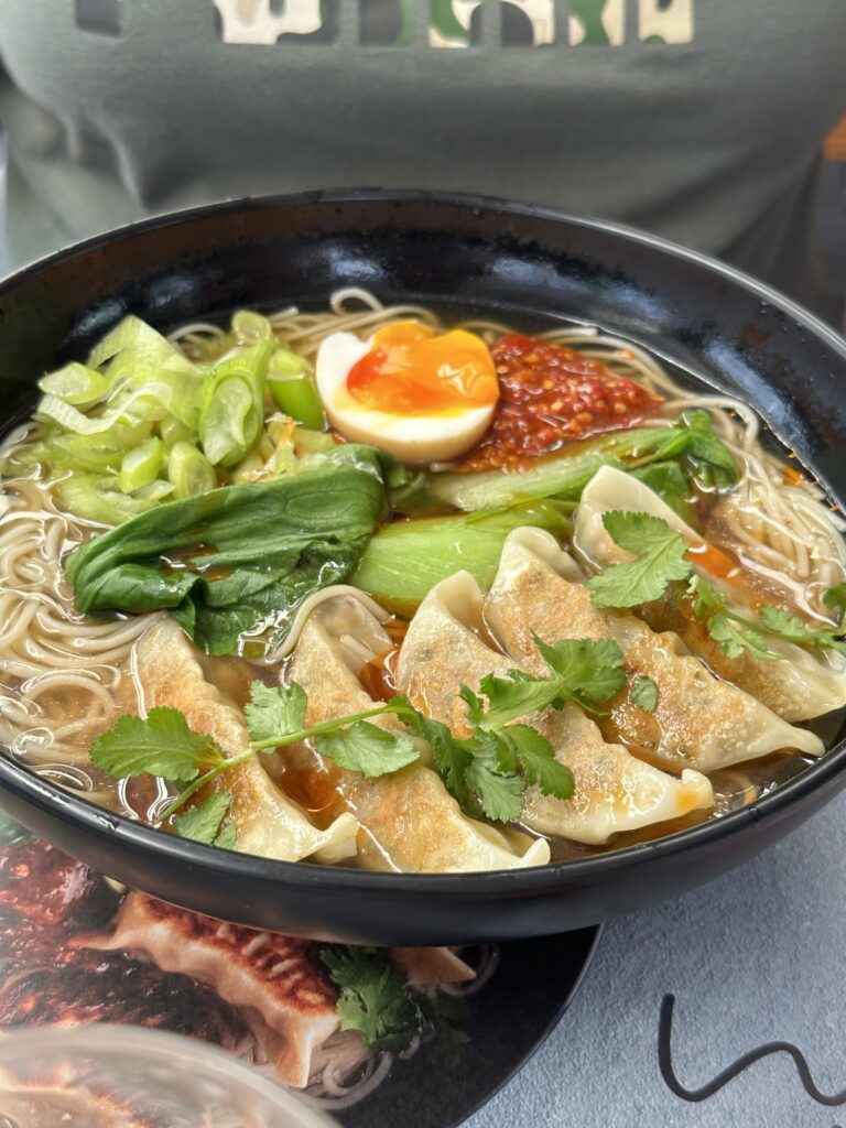 Bowl of ramen with dumplings, soft-boiled egg, greens, and chili paste in broth.