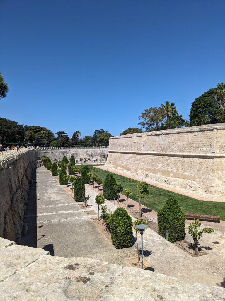 Wide stone fort walls surrounding a landscaped moat with trimmed trees, pathways, and grass under a clear blue sky.