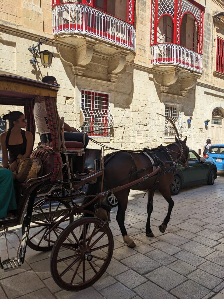 Horse-drawn carriage traveling along a stone street past a beige building with red balconies; passengers seated inside.