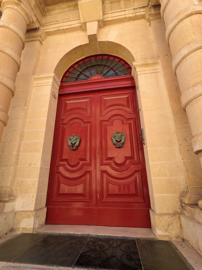 Red double doors with decorative panels and lion-head knockers set in a beige stone archway.