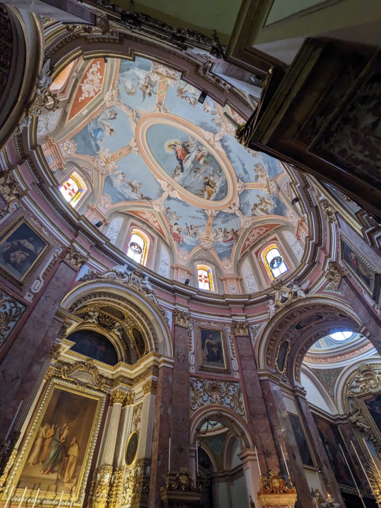 Ornate church interior with frescoed dome ceiling, arched columns, and gilded altars and paintings.