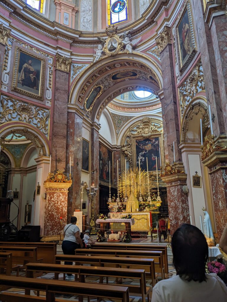 Church interior with wooden pews facing an ornate altar, gilded decorations, paintings, and tall columns; a few visitors seated and standing.