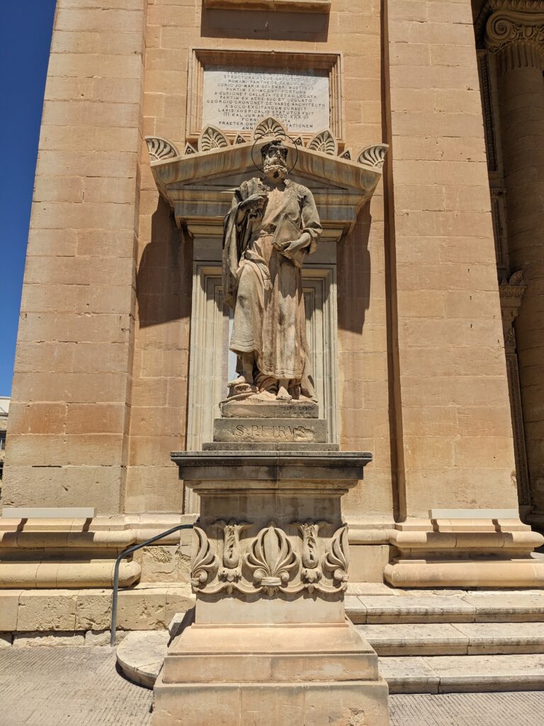 Stone statue of a robed figure holding a book, set in a niche on a beige church façade above a decorative pedestal.