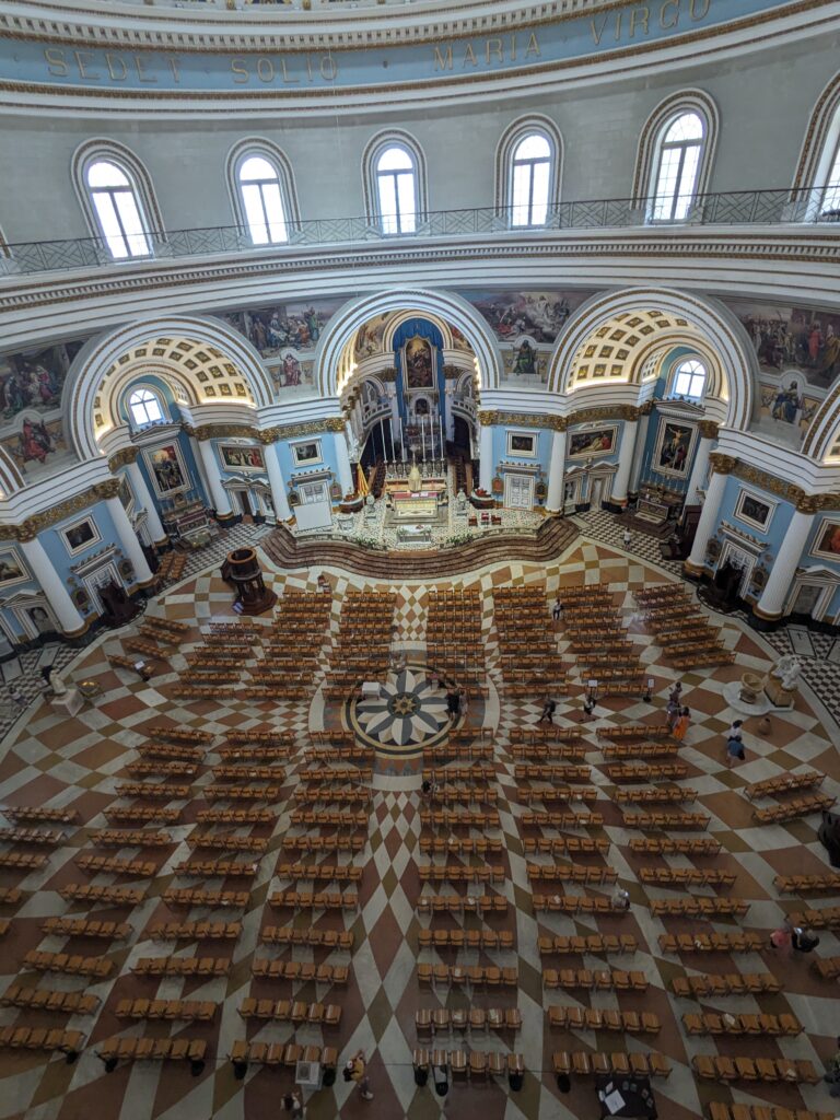 Wide view of a large church interior with rows of wooden chairs arranged on a patterned floor facing an ornate altar beneath arched ceilings.