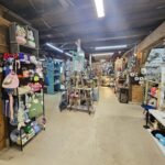 A cozy craft store interior with shelves and displays full of handmade items, yarn, hats, bags, and decorative crafts. The space has wooden beams, bright lights, and a rustic, inviting atmosphere.