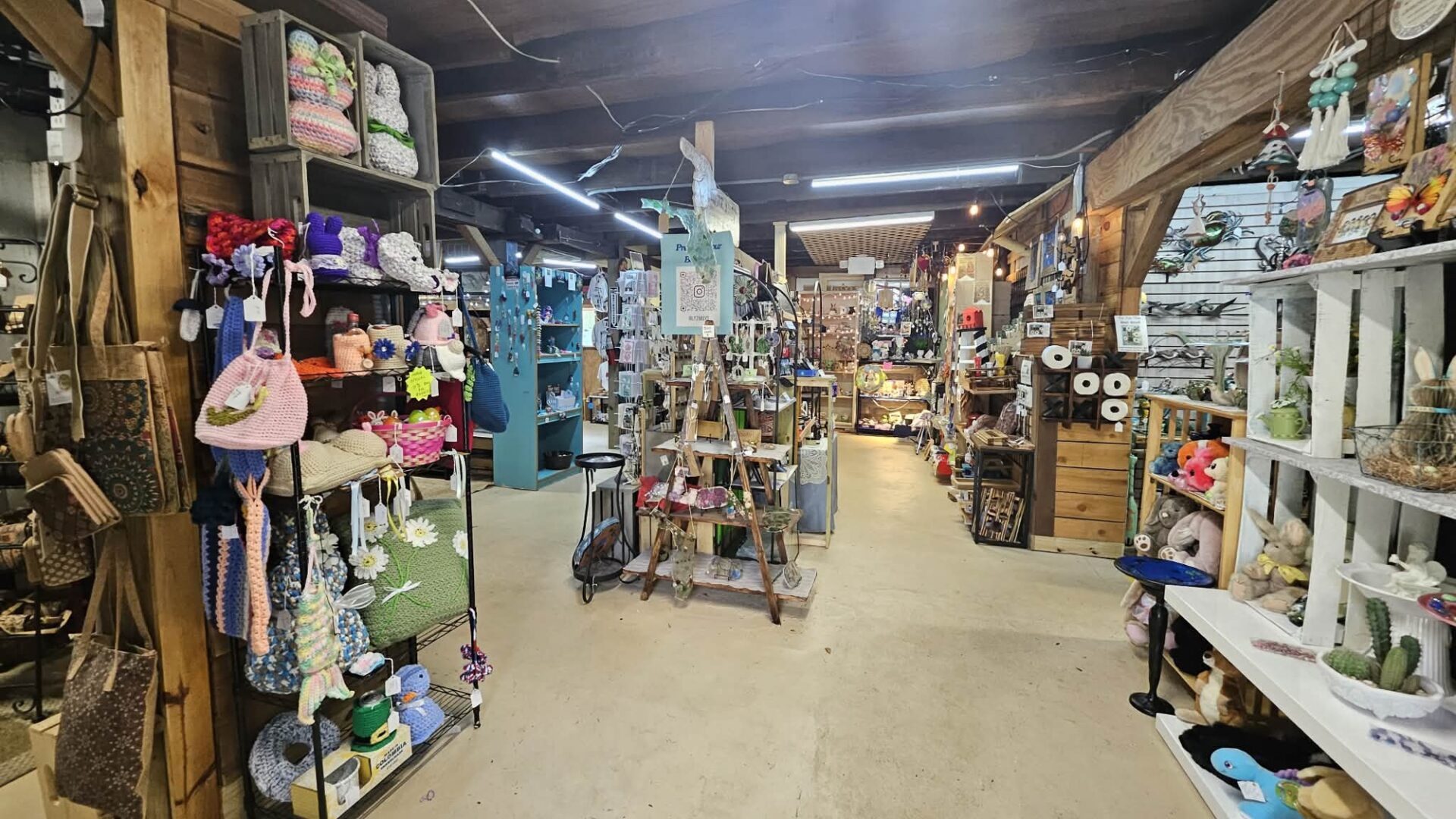 A cozy craft store interior with shelves and displays full of handmade items, yarn, hats, bags, and decorative crafts. The space has wooden beams, bright lights, and a rustic, inviting atmosphere.