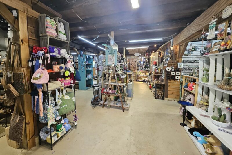 A cozy craft store interior with shelves and displays full of handmade items, yarn, hats, bags, and decorative crafts. The space has wooden beams, bright lights, and a rustic, inviting atmosphere.