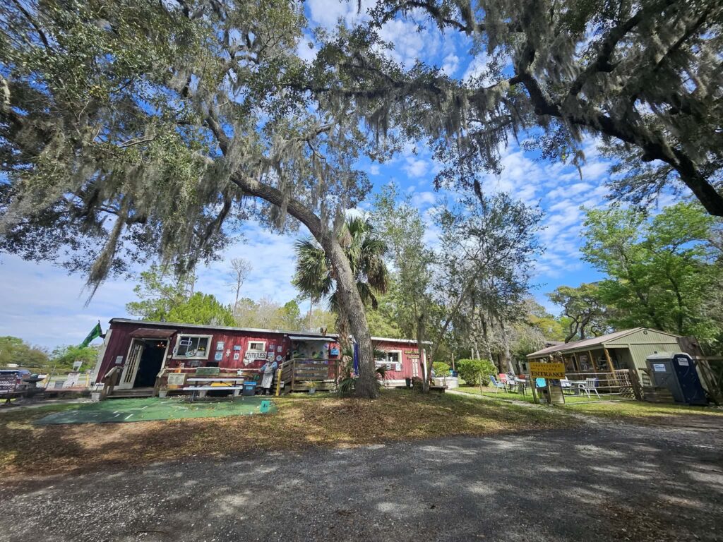 A rustic red building with a porch sits under large oak trees draped with Spanish moss. Bright daylight, scattered clouds, green grass, and a small shed and portable toilet are visible on the right side.