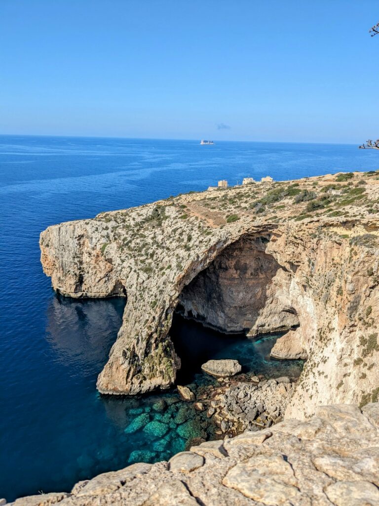 Rocky coastal cliffs with a large sea cave and clear blue water below.