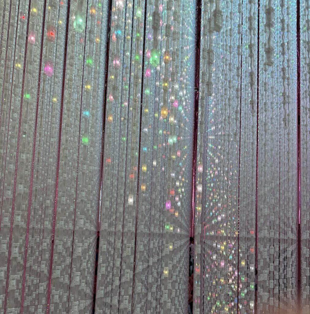 Rows of vertical strings with small, multicolored LED lights create a shimmering, glowing effect in a dimly lit space, giving a sense of depth and a dreamy, immersive atmosphere.