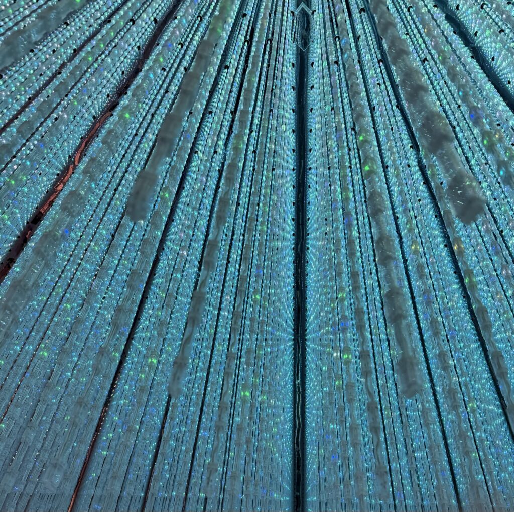 Close-up of a blue and turquoise patterned surface, featuring radiating lines and small dots that shimmer with a holographic, iridescent effect, creating a visually striking and abstract geometric design.