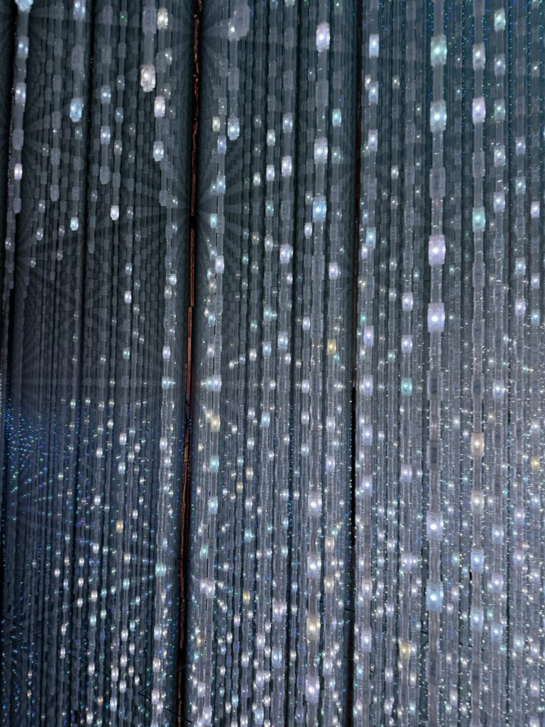 Rows of hanging LED light strands create a sparkling, immersive effect, with glowing white and blue lights suspended in a dark, reflective space.