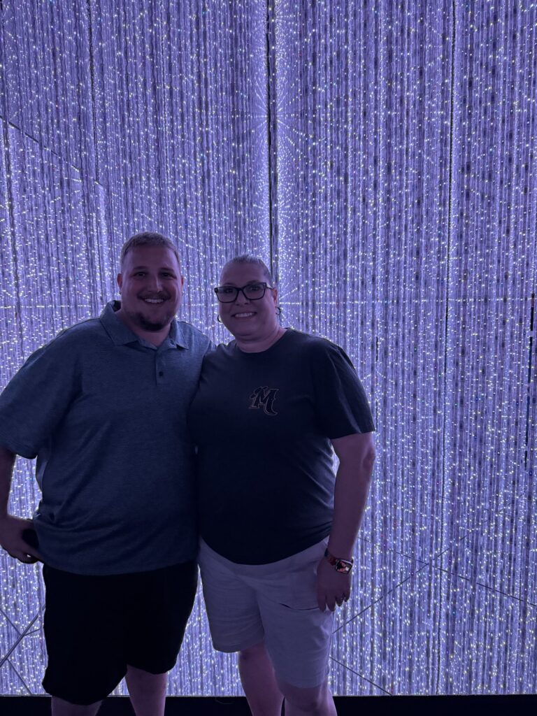 Two people stand smiling together in front of a backdrop of vertical strands of glowing lights, creating a sparkling, immersive atmosphere. Both wear casual clothing and glasses.