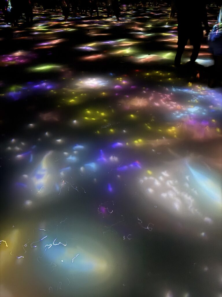 Colorful lights are reflected on a glossy surface in a dark room, with several shadowy figures standing or walking in the background. The scene has a vibrant, dreamy, and abstract atmosphere.