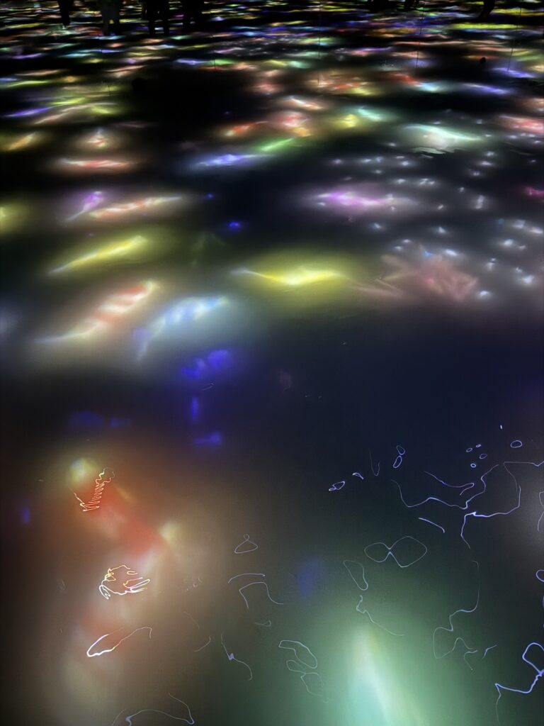 Colorful lights are reflected on the surface of water in a dark setting; abstract patterns and ripples are visible, with some silhouettes of people faintly seen at the top of the image.