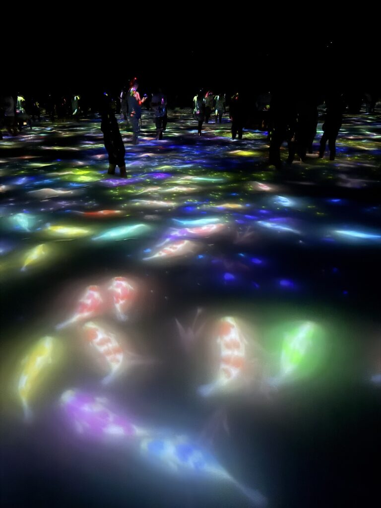People walk on a floor illuminated with colorful, glowing digital images of koi fish and light patterns, creating an immersive, interactive art experience in a dark space.