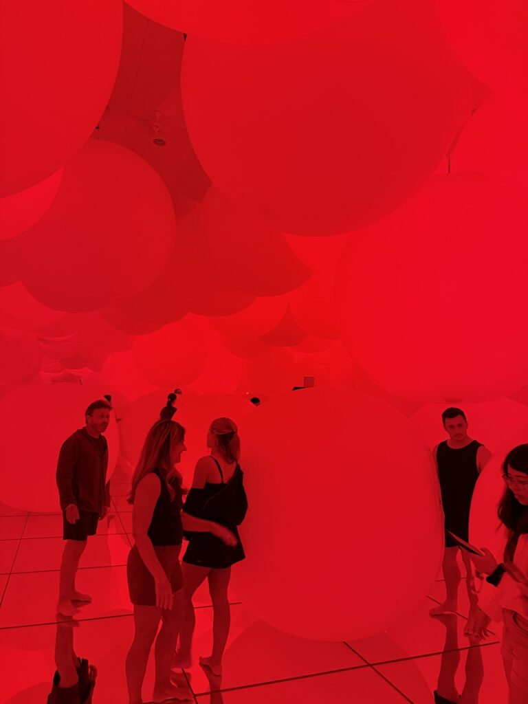 A group of people stand in a room filled with large, round, red balloons hanging from the ceiling and floating on the floor, all bathed in intense red lighting.