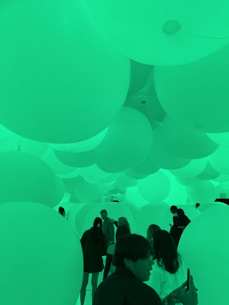 People walking among large, inflated white balloons in a room lit with green light. Some are taking photos while others look around, surrounded by the oversized floating spheres.