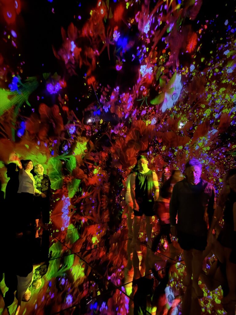 People walk through a dark room filled with vibrant, multi-colored lights and floral patterns projected on the walls and floor, creating an immersive and surreal atmosphere.