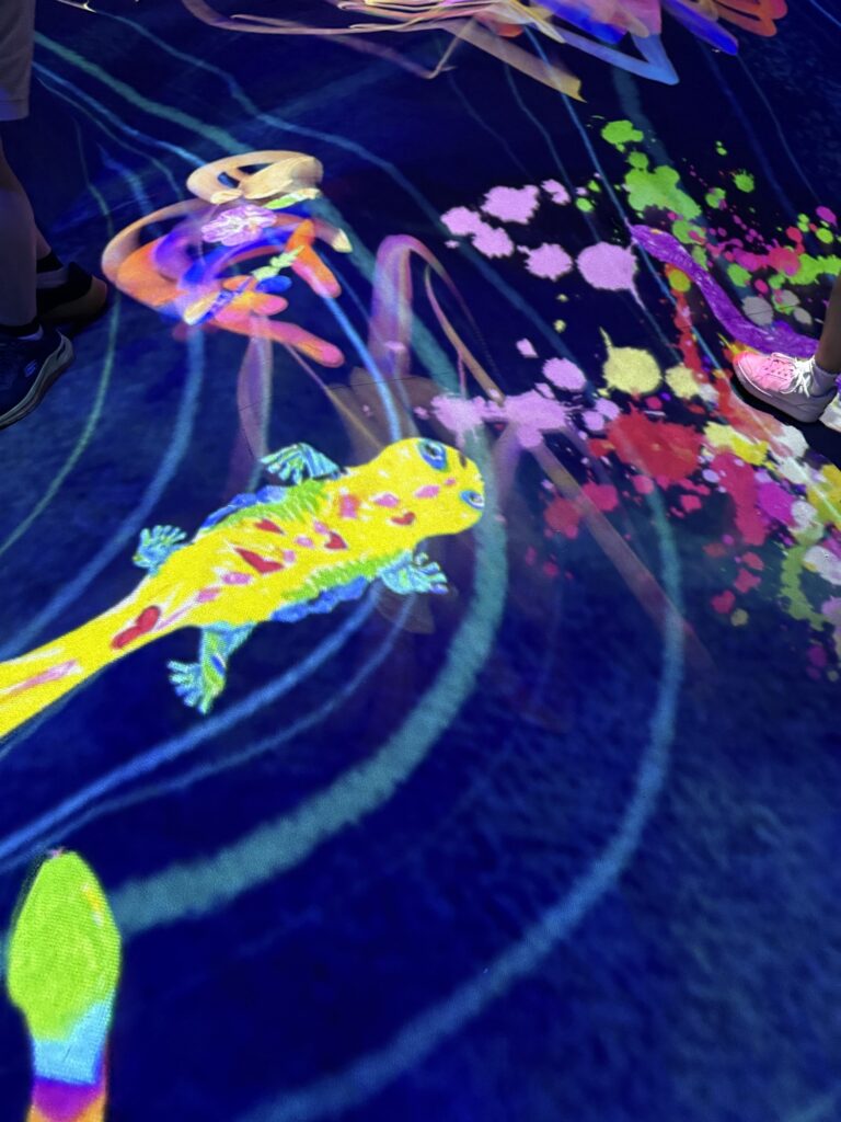 Colorful digital projection of a salamander and abstract shapes on a dark floor, with splashes of bright paint and several people’s legs and shoes visible around the artwork.