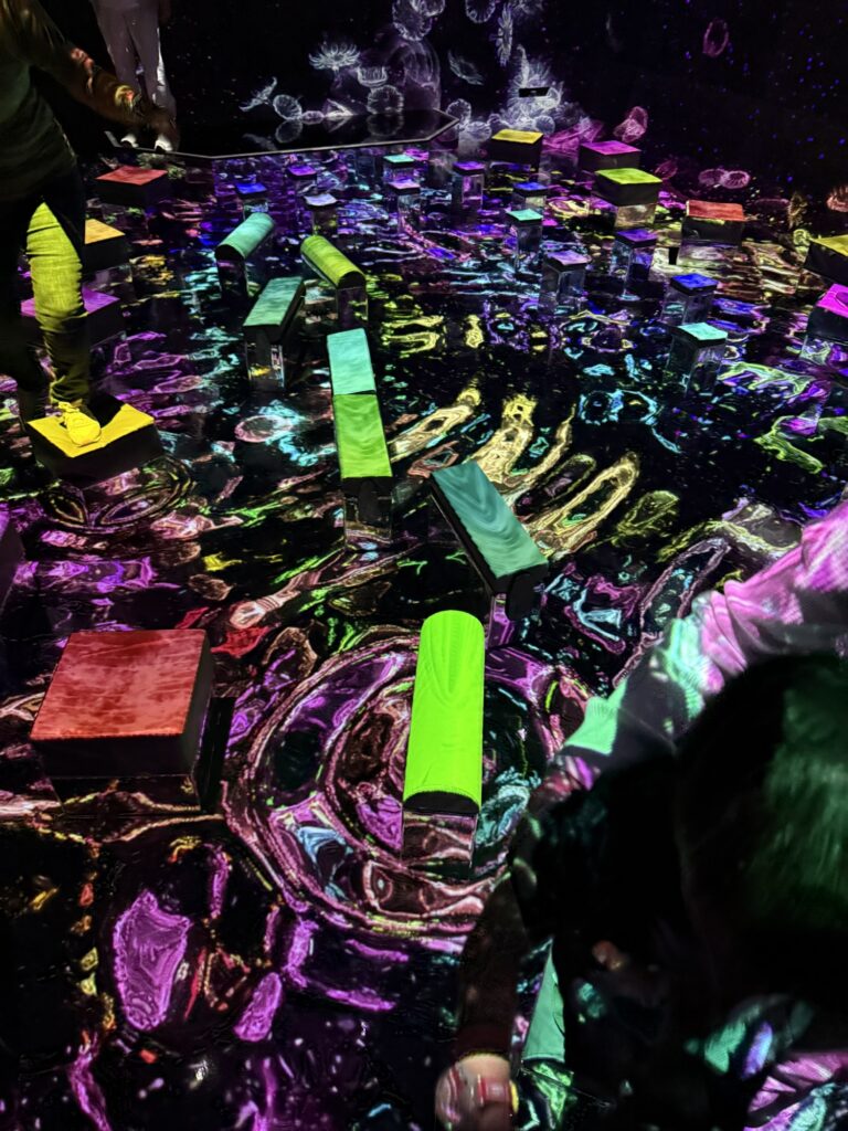 People walk on glowing, rectangular blocks arranged on a floor illuminated with colorful, swirling, abstract light patterns in a dark, immersive art installation.