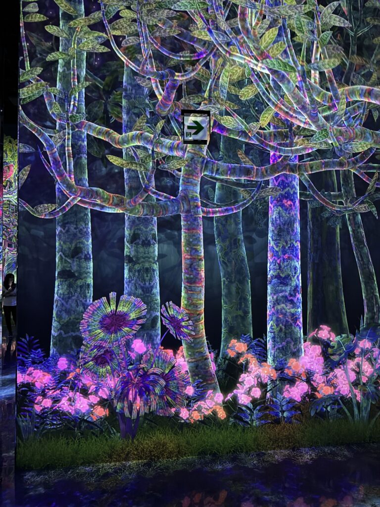 A glowing, neon forest scene with tall, colorful trees and vibrant plants illuminated under blacklight. An exit sign with a right arrow hangs above the scene.